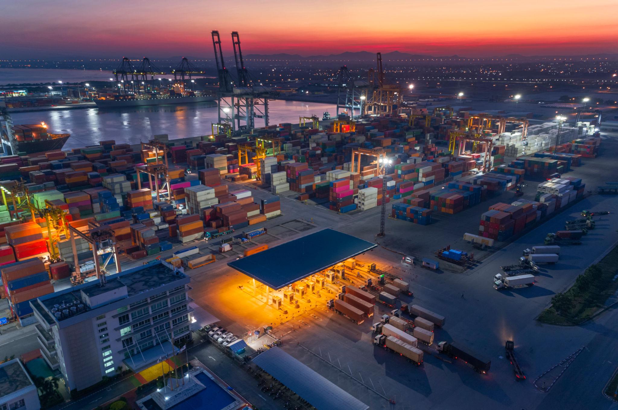 Container port at sunset with stacked containers and cranes silhouetted against a dusk sky, global trade infrastructure